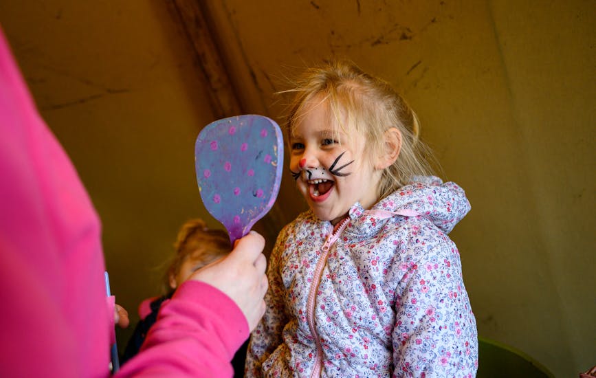 A child smiles into a handheld mirror at her Easter bunny face paint