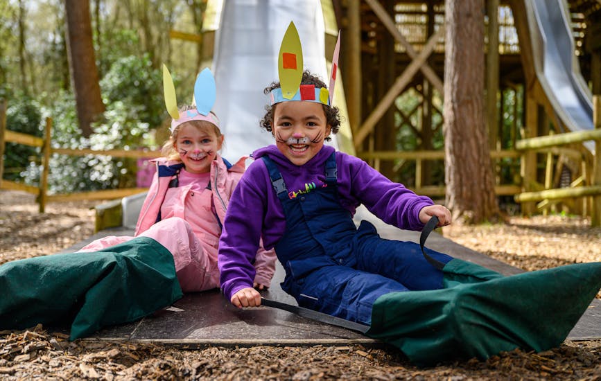 Two children in Easter bunny face paint and headbands smile as they slide down the Slippery Slopes at BeWILDerwood