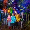 A family walk in a brightly lit woodland filled with colourful lights during BeWILDerwood's Glorious Glowing Lantern Parade